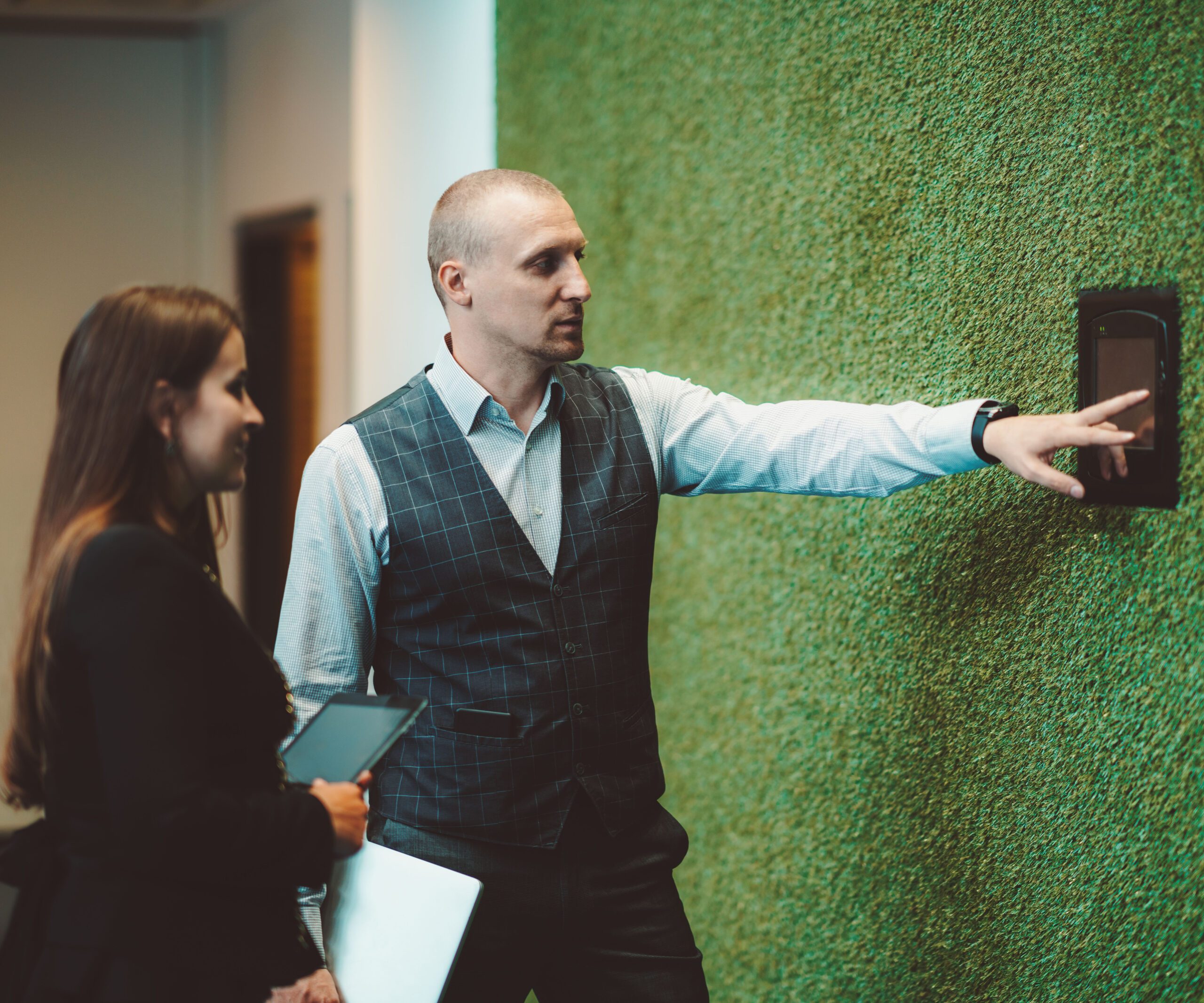 A caucasian businessman in a plaid vest is showing his female colleague how to use indoor climate control and office light illumination terminal placed on the wall with green artificial grass covering